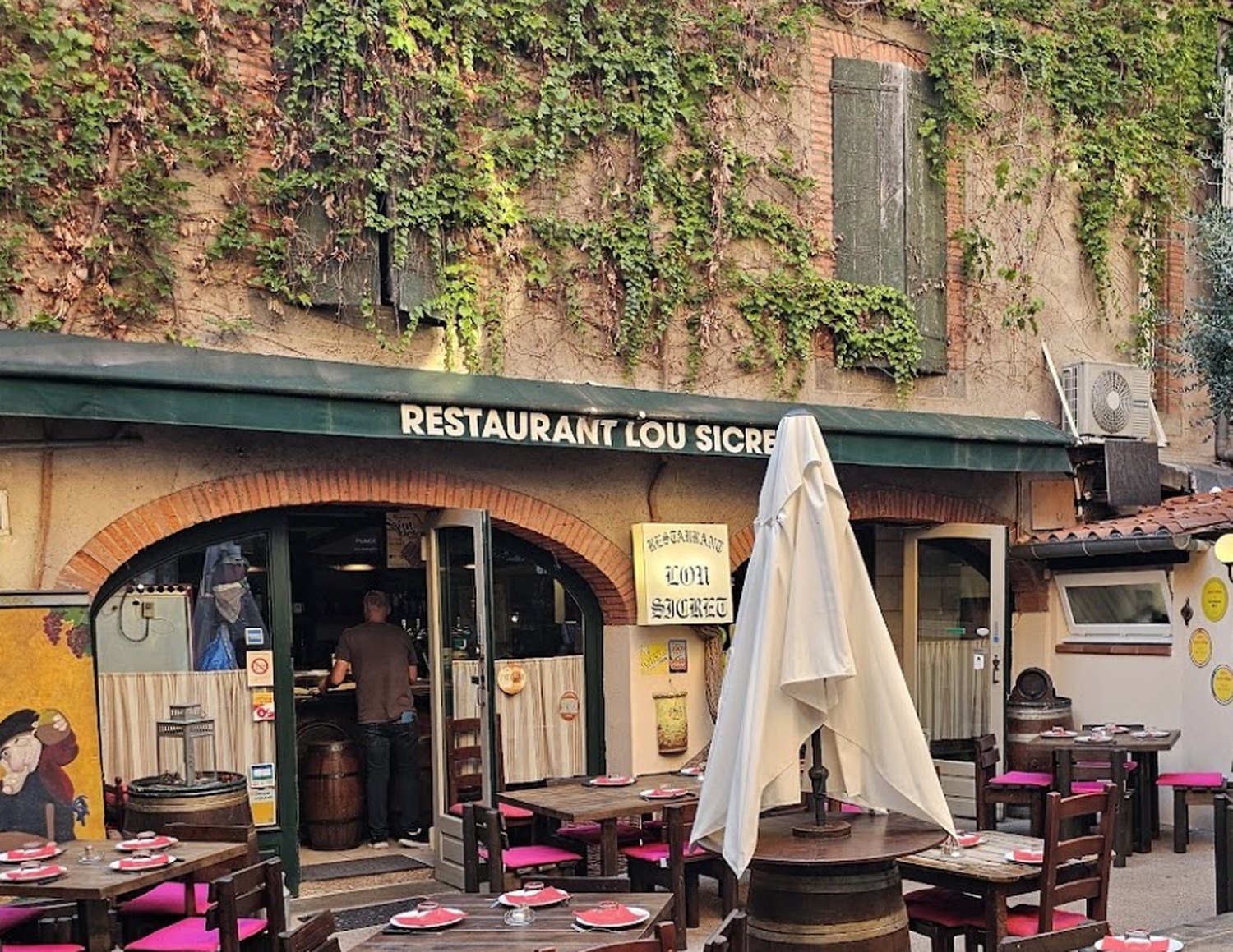 La cour de Lou Sicret à Albi : mur de brique couvert de vigne, store vert « Restaurant Lou Sicret », arche de brique et tables aux coussins fuchsia.