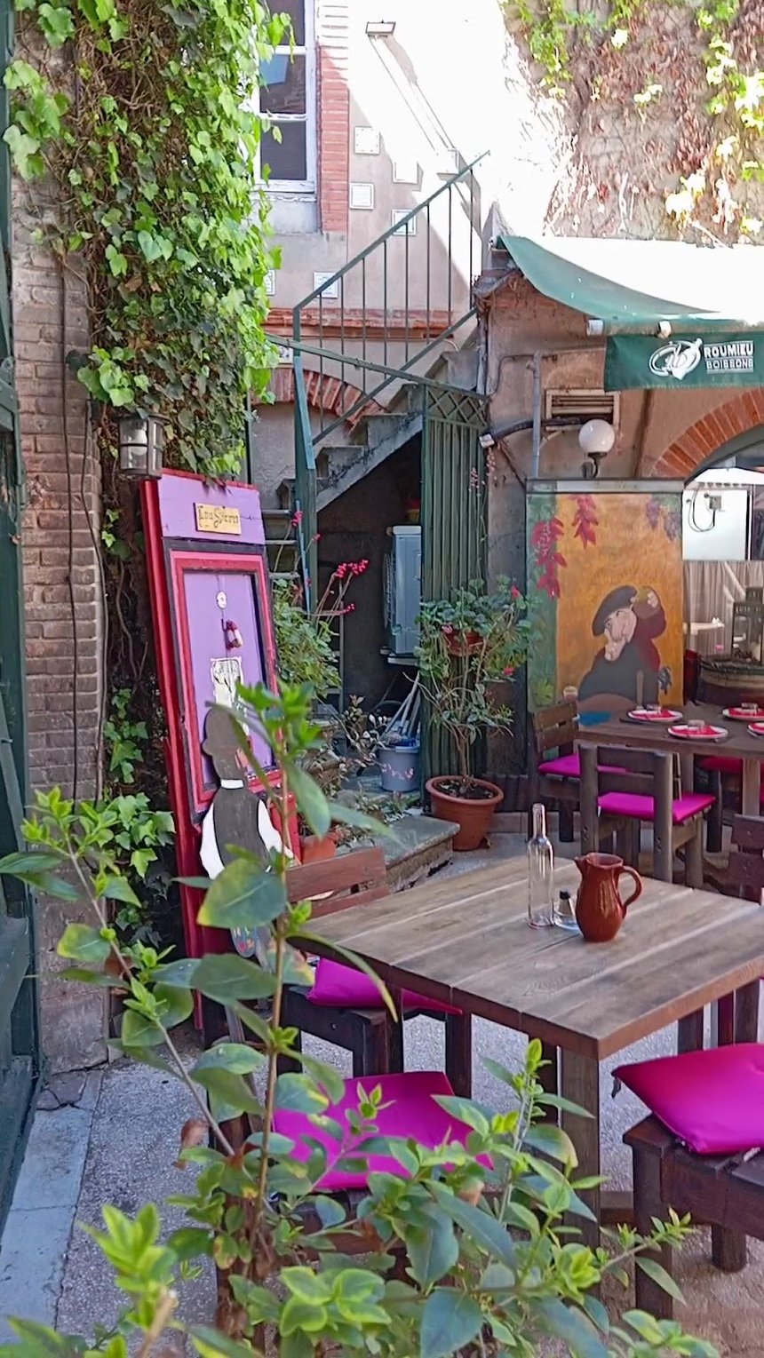L’impasse qui mène à la cour : brique, plantes en pot, tables et coussins fuchsia.