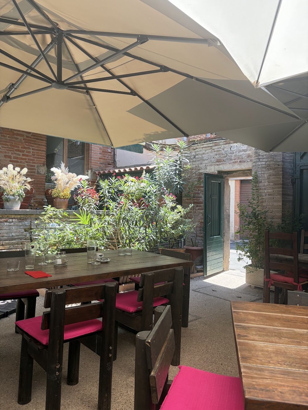 La cour sous les grands parasols, murs de brique, tables en bois et coussins fuchsia.