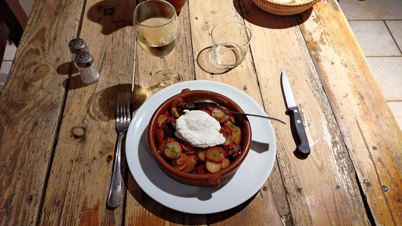Une table en bois brut de la cour, vue de dessus : cassole, couverts, verre de vin blanc.
